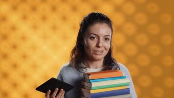 Woman debating between physical books and ebooks on ereader, isolated over studio background. Bookworm comparing paper novels and digital literature on tablet, technology concept, camera A photo