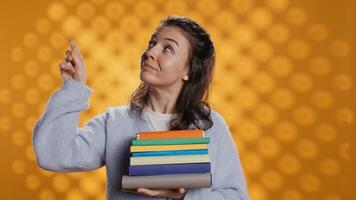 Portrait of smiling woman pointing towards empty copy spaces while holding pile of books. Radiant person with stack of novels in arms promoting reading, showing messages, studio background, camera B photo