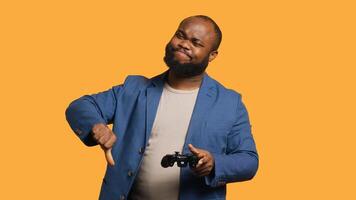 Upset man showing thumbs down sign gesturing holding controller, disapproving of videogames. Displeased BIPOC person against gaming, doing rejection hand gesture, studio background, camera A photo
