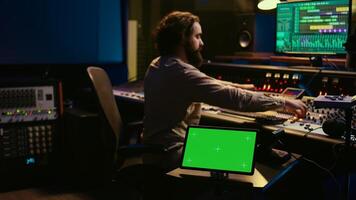 Sound designer processing and recording tracks in control room, using mixing console and sliders to adjust volume level. Technician working with greenscreen tablet in professional studio. Camera B. photo