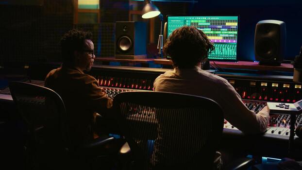 Diverse team collaborating on creating a new hit song in control room, mixing and mastering sounds in post production. Artist and audio expert editing volume level and sound settings. Camera B. photo