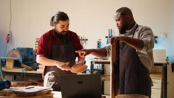 Woodworker looking over blueprints on laptop, brainstorming with colleague next steps in wood processing. Manufacturer and BIPOC apprentice reading furniture assembly schematics in studio, camera B photo