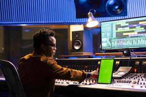 Sound technician mixing tracks next to greenscreen tablet in control room, operating panel board knobs and faders. African american producer recording hit song and processing sounds. photo