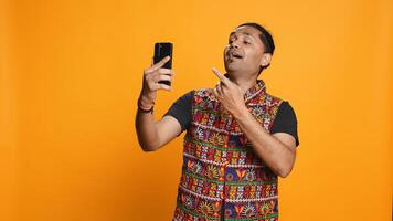 Joyous Indian man greeting friend over online videocall on cellphone, doing friendly gestures. Excited person bonding with mate using mobile phone videoconference, studio background, camera B photo