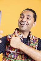 Happy man using cellphone to take selfies, doing thumbs up gesture. Jolly social media user taking photos using phone selfie camera, doing like hand gesturing, studio background