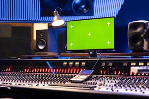 Mockup display on computer next to control desk soundboard, empty music recording studio with professional equipment and gear. Panel board with faders and knobs for post production. photo