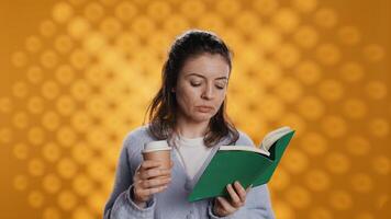 Woman reading book, turning page and holding disposable cup of coffee, isolated over studio background. Bookworm enjoying novel and caffeinated drink early in the morning, camera B photo
