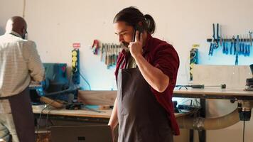 Cabinetmaker receiving telephone call from client inquiring about furniture assembling commission progress. Carpenter in assembly shop talking on smartphone with customer, camera A photo