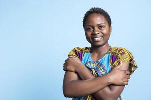 Portrait of optimistic african american girl standing on camera while dressed in ancestral vibrant outfit on blue background. Gorgeous woman, young and ecstatic posing in the studio. photo