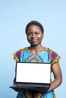 African american model creates ad commercial with white display on pc, pointing at portable computer that shows blank copyspace display. Young adult smiling at camera presenting laptop. photo