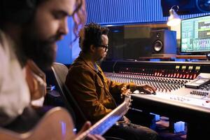 Talented singer composer recording guitar notes in professional music studio, using electro acoustic sounds to add on record. Artist working with audio technician in control room, post production. photo