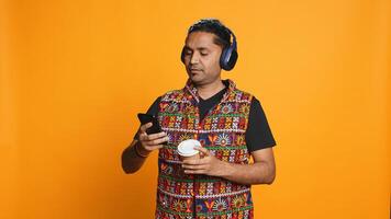 Joyous man listening music, having fun dancing, enjoying hot beverage. Upbeat person wearing headphones, doing funky movements, drinking coffee, studio background, camera B photo