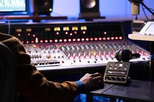 African american expert working at mixing panel in recording studio. Using control desk soundboard and digital audio software interface on pc, motorized faders and switchers to edit tracks. photo