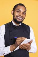 Friendly restaurant waiter taking notes of meal orders for clients, using information on the notepad for the bill. Butler serving people for customer satisfaction against yellow background. photo