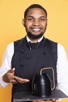 Friendly smiling butler carrying a teapot to serve clients with tea at the table, five star restaurant services. Waiter in uniform focuses on customer satisfaction,catering industry concept. photo