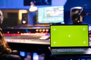 Focused audio engineer recording tracks on control panel board next to laptop with greenscreen, adjusting volume levels on mixing console. Sound designer creating music for his new album. photo