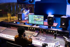 African american audio expert editing tunes with digital software on pc, mixing and mastering tracks with motorized faders and switchers. Professional music producer working on new songs. photo