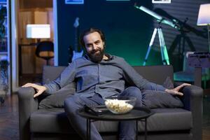 Portrait of happy person using TV to binge series on subscription based streaming services, enjoying bowl of popcorn. Joyful man relaxing at home watching VOD shows on television display, eating snack photo