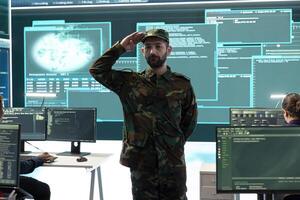 Army soldier showing the respectful military salute in government high tech agency, hackers in hybrid warfare strategies. People working with modern software and technology to secure information. photo