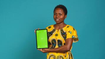 Feminine person in native clothing holds a tablet with mockup screen, creating an advertisement against blue background in studio. Young woman smiling and presenting copy space. Camera A. photo