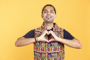 Portrait of smiling caring indian man doing heart symbol shape gesture with hands, showing kindness. Joyous affectionate person doing tender love gesturing, isolated over studio background photo