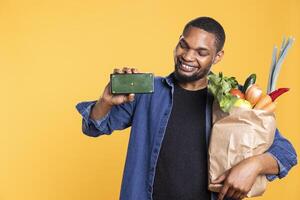 Happy charismatic man presenting greenscreen layout on camera, carrying a bag of groceries and smiling. Confident vegan person showing isolated mockup template on phone display. photo