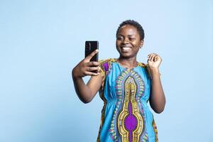 African american person having fun with photographs on smartphone, smiling at the camera and taking pictures in studio. Optimistic positive woman enjoy taking photos in ethnic attire.