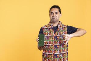 Upset indian man shows thumbs down sign gesturing holding isolated screen phone, disapproving. Person does rejection hand gesture regarding mockup smartphone, studio background photo