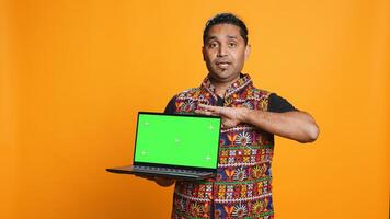 Upbeat tech specialist doing promotion using isolated screen laptop, studio background. Cheerful indian person holding chroma key notebook, doing advertisement for it, camera B photo