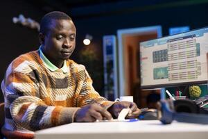 Self employed black man writing monetary data on an adhesive note, creating a post it with international capital funds options for expansion. Expert validates budget balance with a reminder. photo