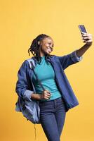 Cheerful woman using smartphone to take selfies and post them on social media. Happy BIPOC person taking photos using phone selfie camera, isolated over yellow studio background
