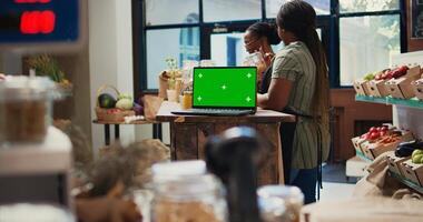 Women chats about products next to laptop with greenscreen, supporting sustainable lifestyle and farming. Buyer looks at pasta in jars, vendor having device with isolated mockup layout. Tripod shot. photo
