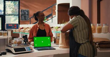 Vendor using a laptop with greenscreen layout at store checkout, serving regular customer with natural products. Women chatting next to pc showing isolated display and mockup copyspace. Tripod shot. photo