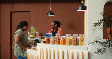 Local vendor putting merchandise on scale at checkout, ensuring the best pricing value for regular client at grocery store. African american retailer serving buyer, eco friendly. Handheld shot. photo