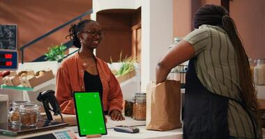 Woman approaching checkout with greenscreen on tablet, buying natural food from local vendor. African american seller using isolated mockup layout on gadget, chatting with customer. Tripod shot. photo