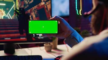 Man in dimly lit apartment watching tutorial on how to finish videogame on green screen smartphone, enjoying day off. Gamer learning online multiplayer shooter tips from on mockup mobile phone photo