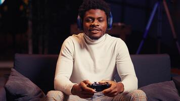 African american man having online gaming session with friends, talking with them through headphones microphone. Gamer playing videogames with teammates on console using gamepad controller photo