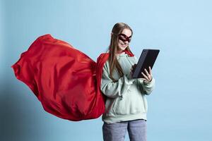 Smiling girl with mask portraying superhero using tablet and wearing red cape, studio background. Happy woman posing as hero in costume with mantle, working on device, being mysterious photo