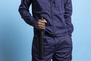 Security agent holding his baton for own safety while on duty, safeguarding and protecting the premises. Trained young bodyguard in work uniform staying alert against blue background. Close up. photo