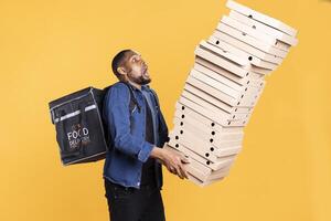 Clumsy african american deliveryman dropping a huge stack of boxes, wasting a lot of pizza and making a mess. Young courier does a mistake and spills a large pile of fast food, reckless accident. photo