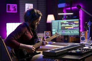 Young artist recording himself playing acoustic guitar in home studio, creating new string accords and editing them on daw software. Musician producing new soundtracks with instrument and amplifier. photo