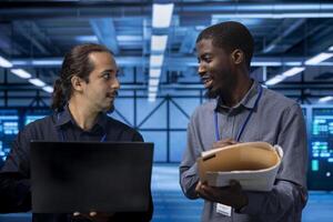 Smiling manager inspecting server room equipment, reading paperwork from folder, talking with employee. Supervisor doing evaluation of data center location, checking files photo