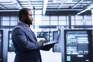 Programmer using laptop in data center with advanced technology, ensuring cybersecurity and efficient network management. Professional in server hub optimizing data center infrastructure with notebook photo
