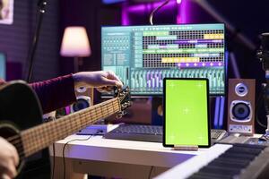 Artist songwriter practicing to play and tune his guitar by twisting knobs, watching online lesson via tablet with greenscreen display. Musician learning acoustic instrument skills in home studio. photo