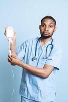 Hospital staff holding IV bag preparing to administer fluids for patients, medications and nutrients via intravenous drip. Man holding recovery tools against blue background in studio. photo