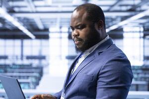 Computer scientist in data center implementing backup solutions to protect against data loss using laptop, ensuring efficiency. African american man in server room resolving technical issues photo