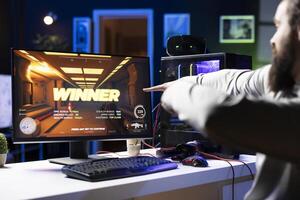 Joyous gamer celebrating after winning esports match during livestream with viewers, pointing to screen. Cheerful man filled with energy after being victorious in videogame photo