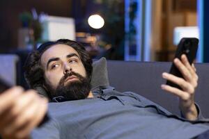 Tired man zapping through channels, watching news broadcast while idling in living room and texting friends. Person using remote control to select TV station to enjoy and using mobile phone photo