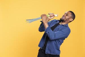 Funny joyful adult using a leek to mimic singing at a microphone, having fun with natural ethically sourced food in studio. African american man fooling around with a green onion. photo