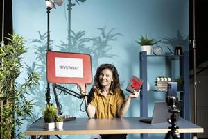Cheerful vlogger advertising giveaway for viewers with cardboard sign, offering gift box, engaging with audience. Upbeat person organizing online contest, explaining rules using copy space board photo